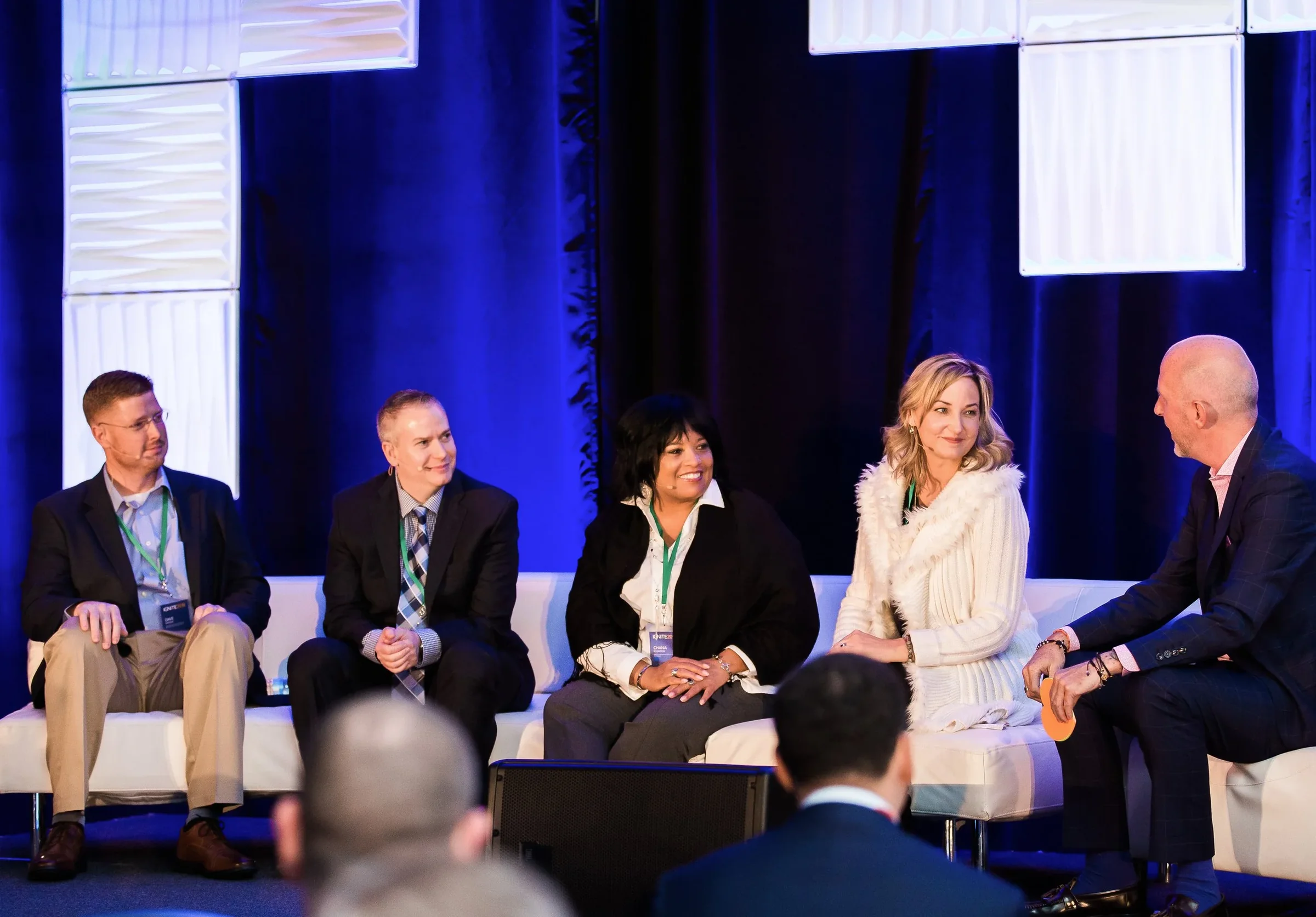 Five panelists in conversation on a blue-lit conference stage.